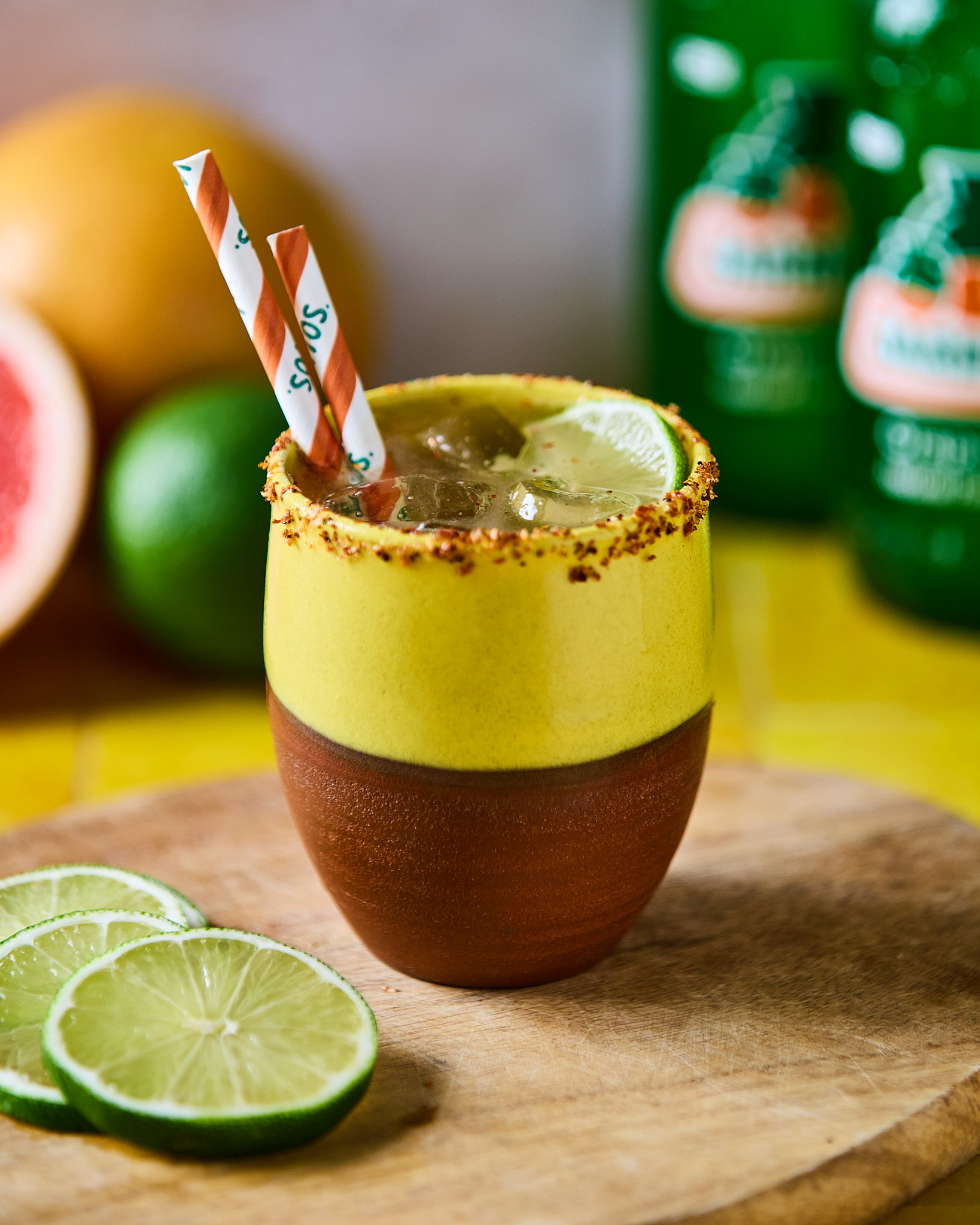 Farverig cocktail med chili-rim, limeskiver og stribet sugerør på træbræt.